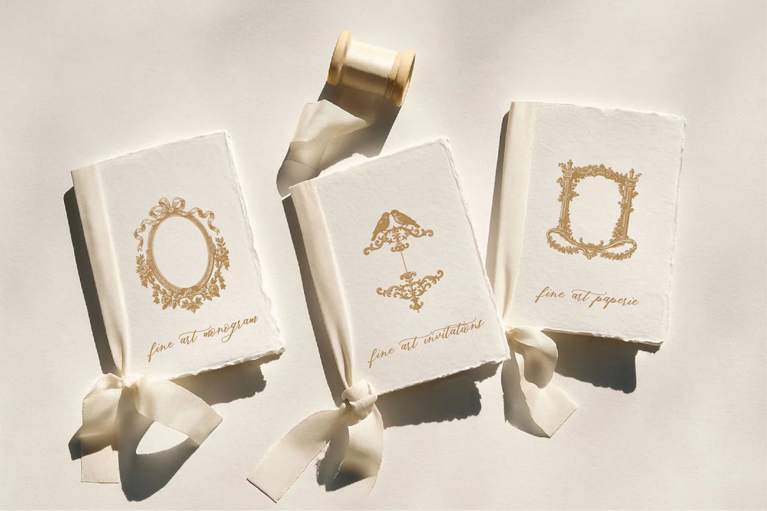 Three fine art wedding monogram vow booklets displayed on handmade deckled-edge paper with silk ribbons, featuring elegant gold crest designs for invitations, monograms, and paperie branding, photographed in natural light on a neutral background.