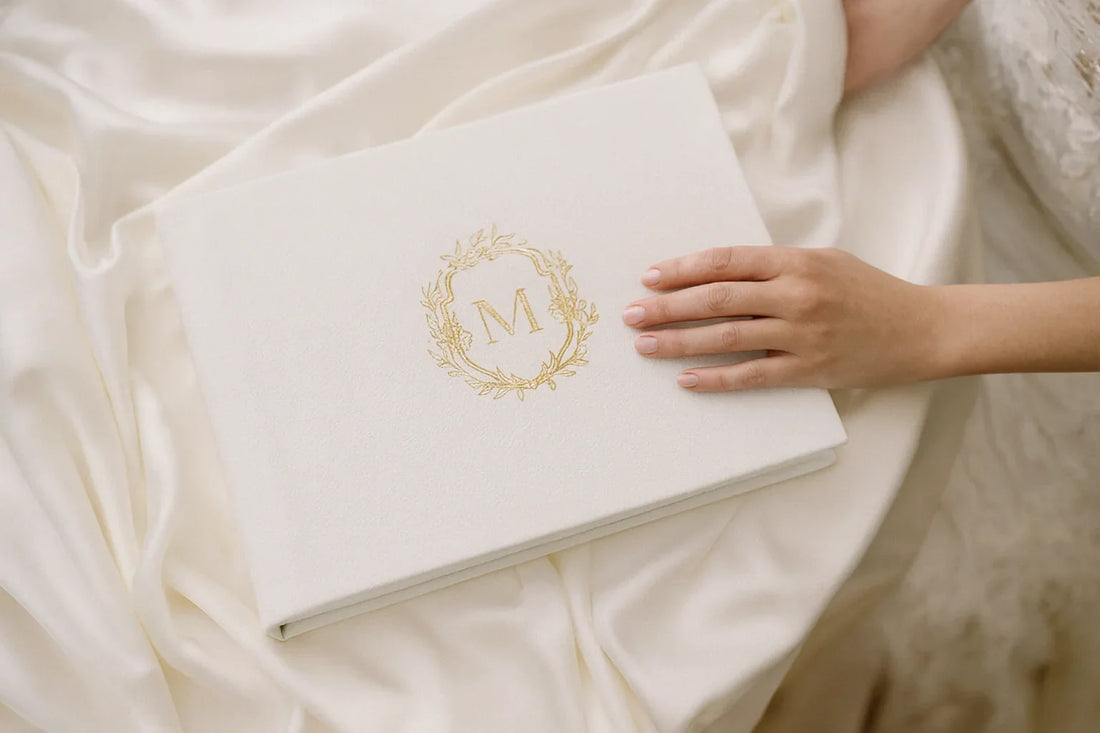 A bride gently touches a white leather wedding photo album engraved with a gold monogram, resting on soft ivory silk fabric — a luxurious heirloom guestbook perfect for elegant fine art weddings.