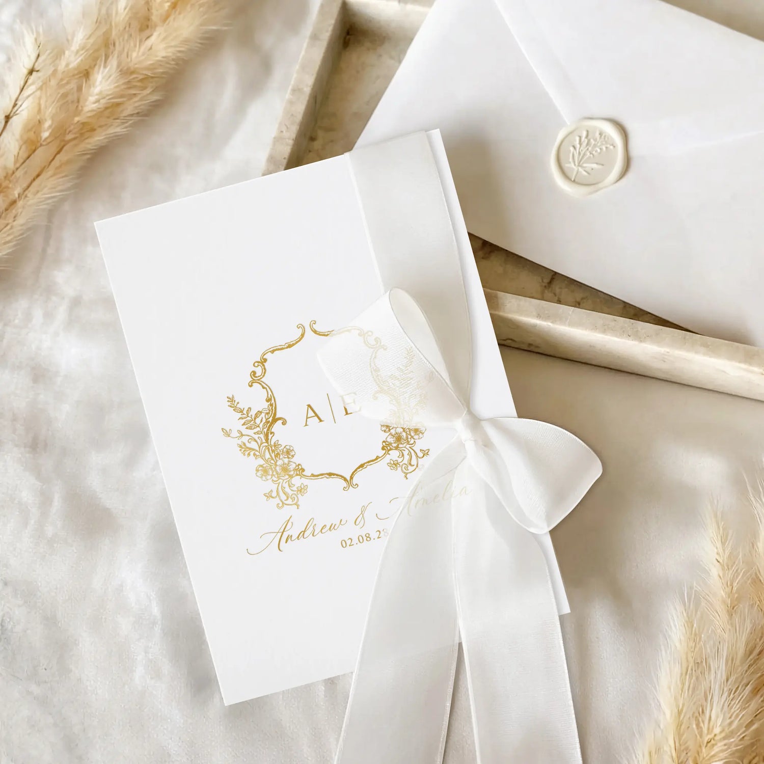 A white wedding invitation card with elegant gold foil monogram and script details, tied with a white satin ribbon, displayed next to a matching white envelope with a wax seal on a neutral fabric background.